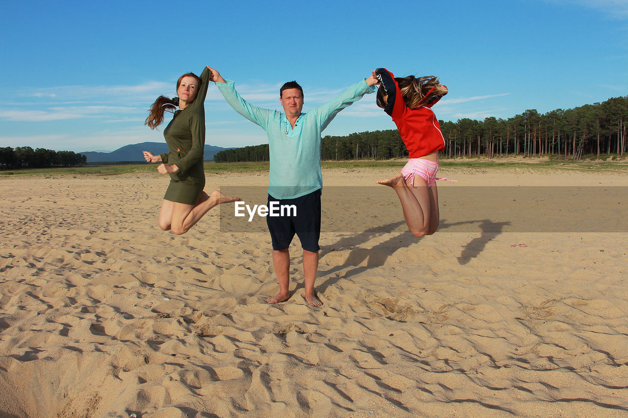 Full length of father holding wife and daughter in hand while standing on beach against sky