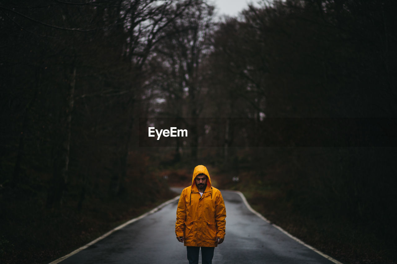 Man in yellow jacket looking down with closed eyes standing on empty asphalt road among green forest