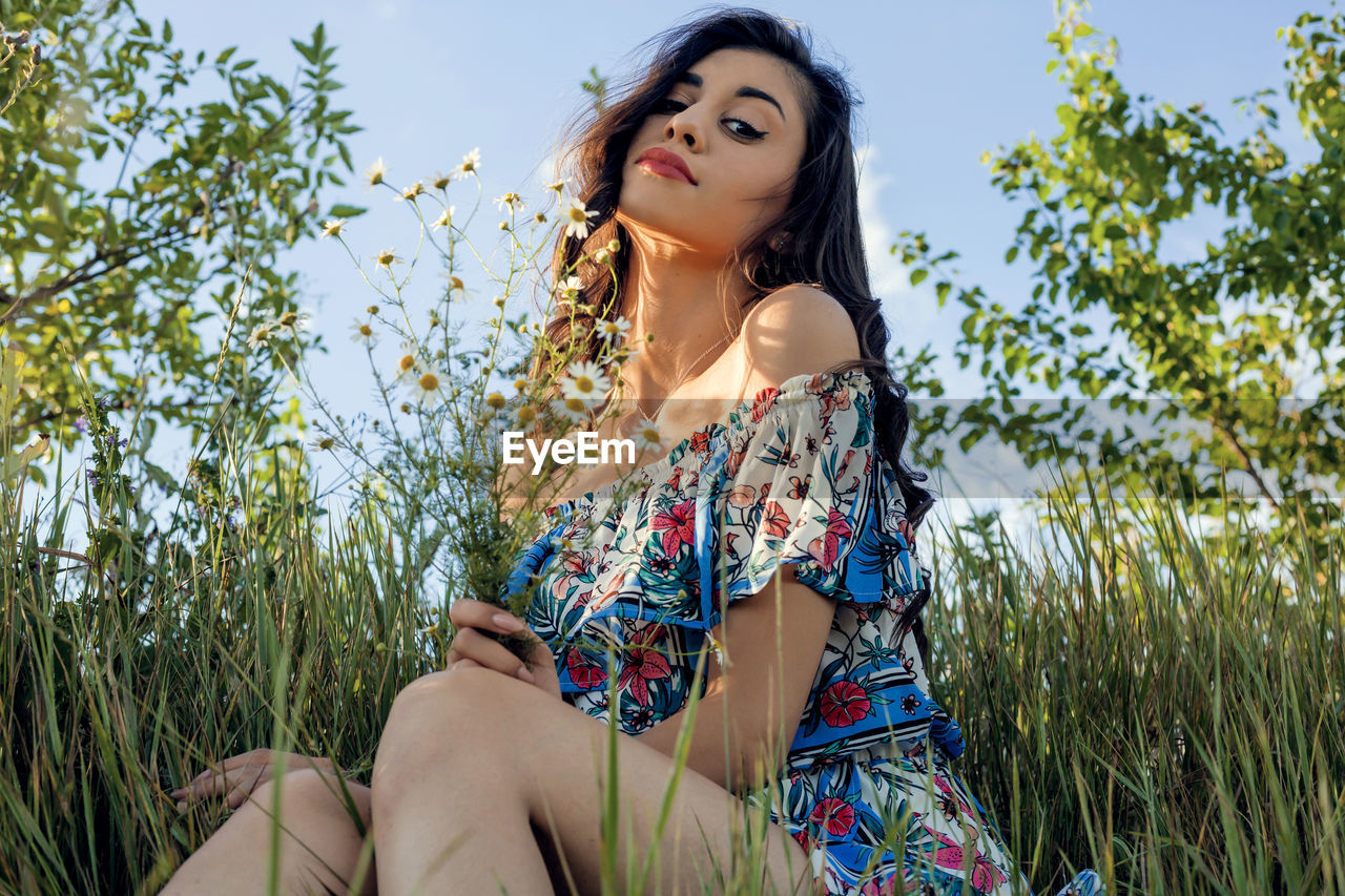 LOW ANGLE VIEW OF WOMAN SITTING ON PLANT AGAINST TREES