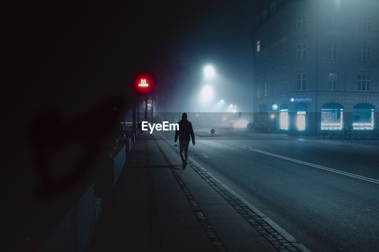 REAR VIEW OF MAN WALKING ON ILLUMINATED STREET AT NIGHT IN CITY