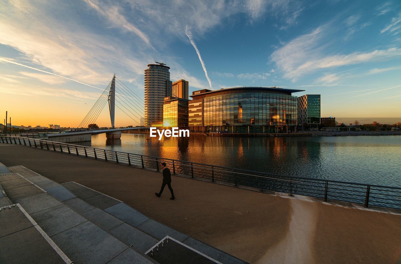 Man walking by river and modern building against sky at sunset