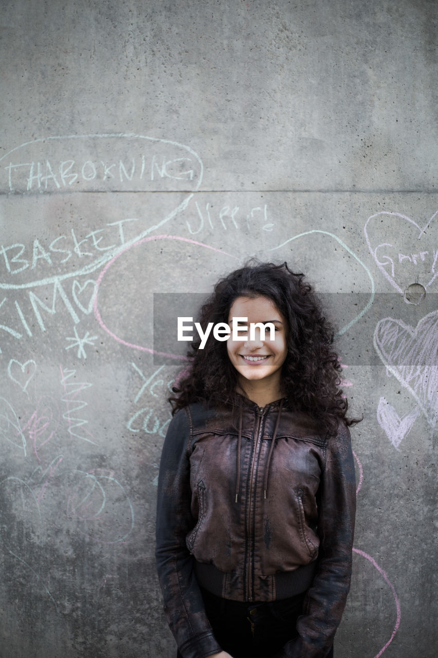 Portrait of smiling teenage girl standing against chalk drawing on gray wall in city