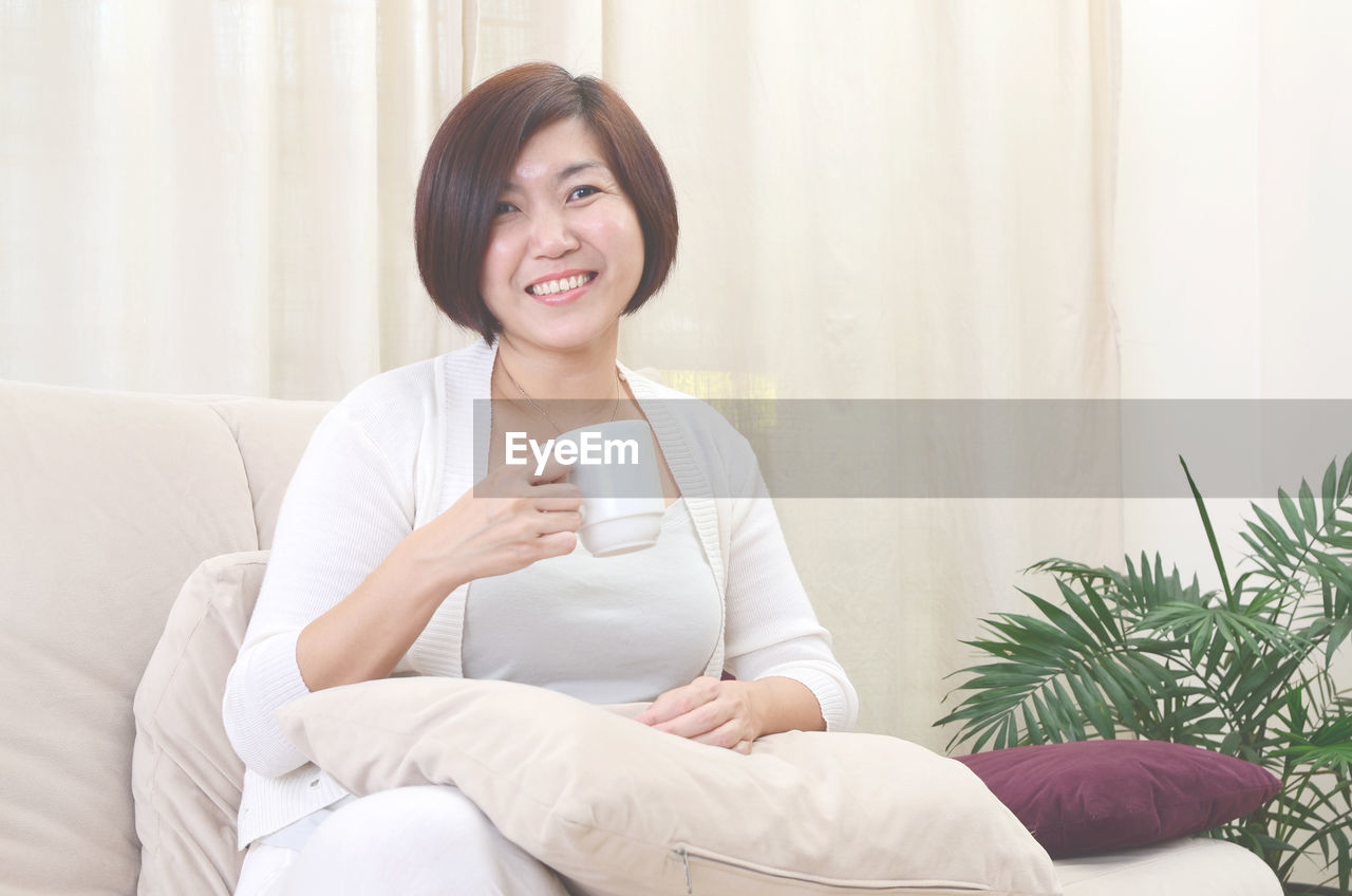 Portrait of smiling woman having coffee while sitting on sofa at home