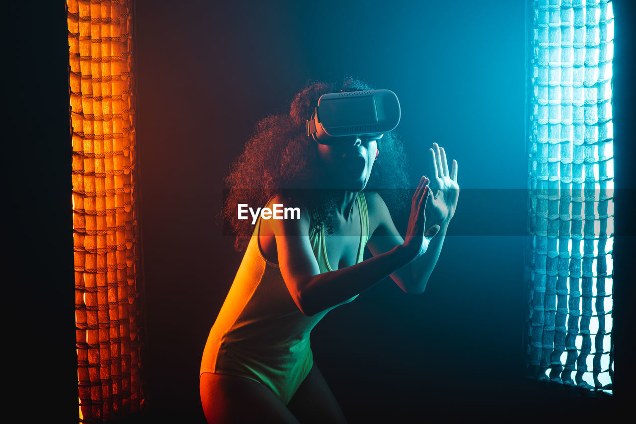 Anonymous astonished ethnic female with open mouth exploring virtual reality in headset on black background