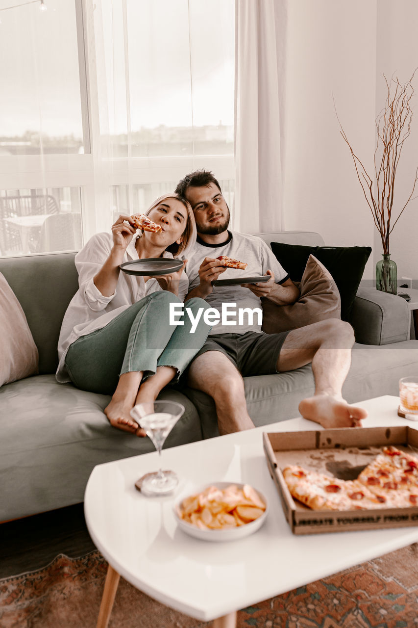 Couple enjoying pizza while sitting on sofa at home