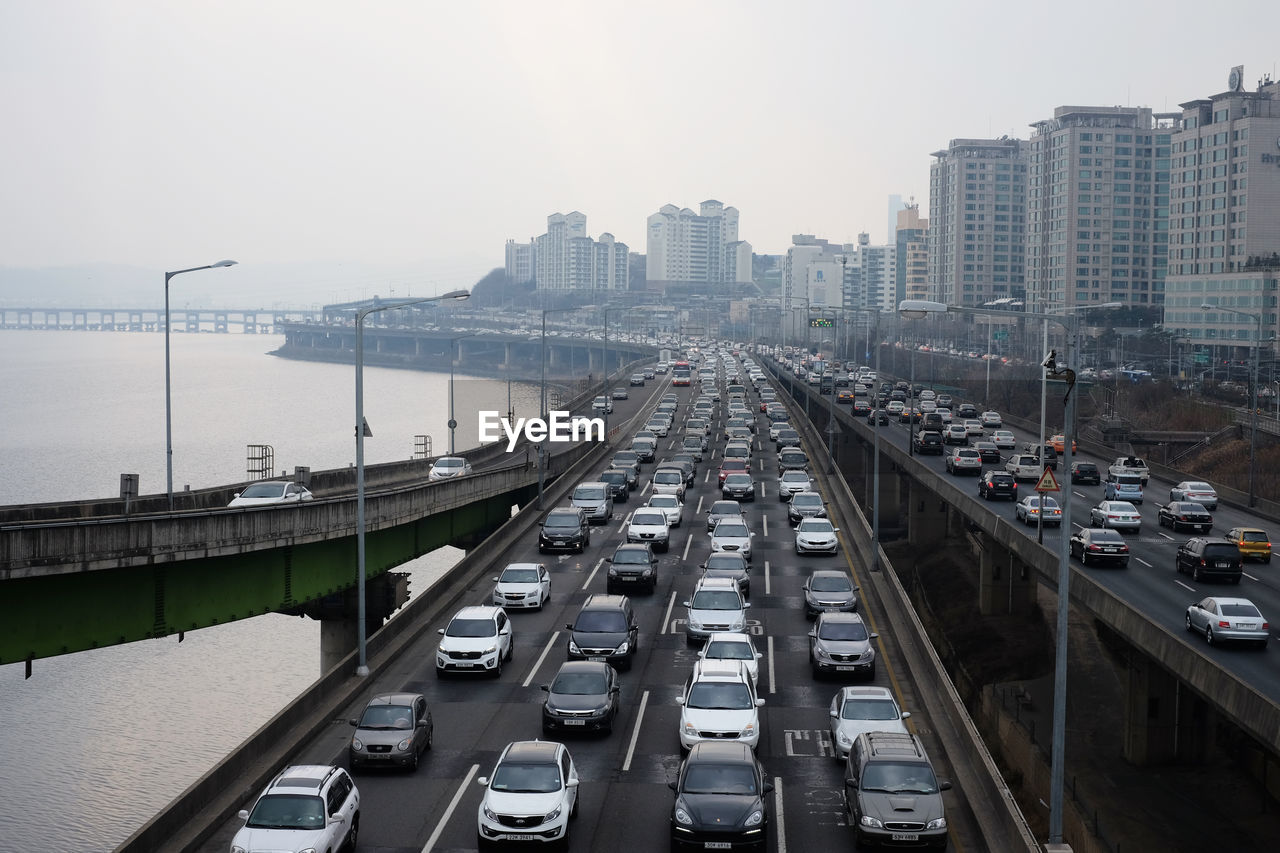 HIGH ANGLE VIEW OF VEHICLES ON ROAD BY BUILDINGS AGAINST SKY