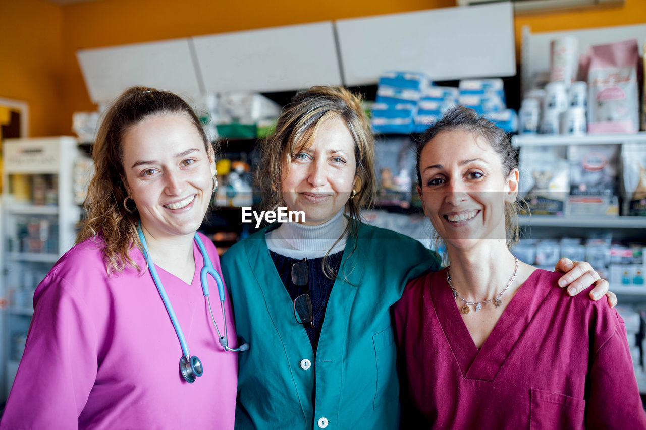 Group of positive female veterinarians in uniforms looking at camera while standing in veterinary clinic with medical supplies during work
