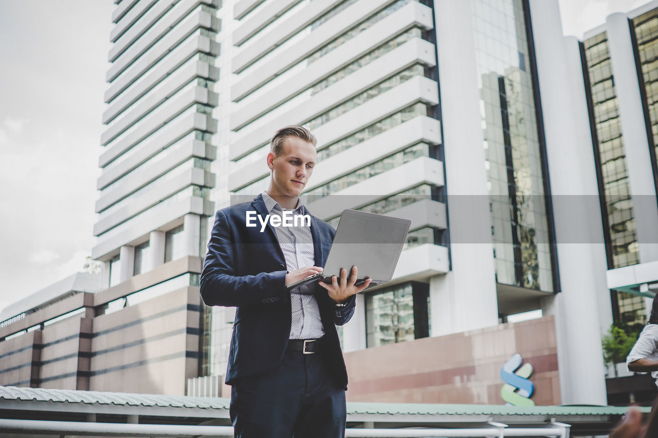 Young businessman using laptop while standing against buildings in city