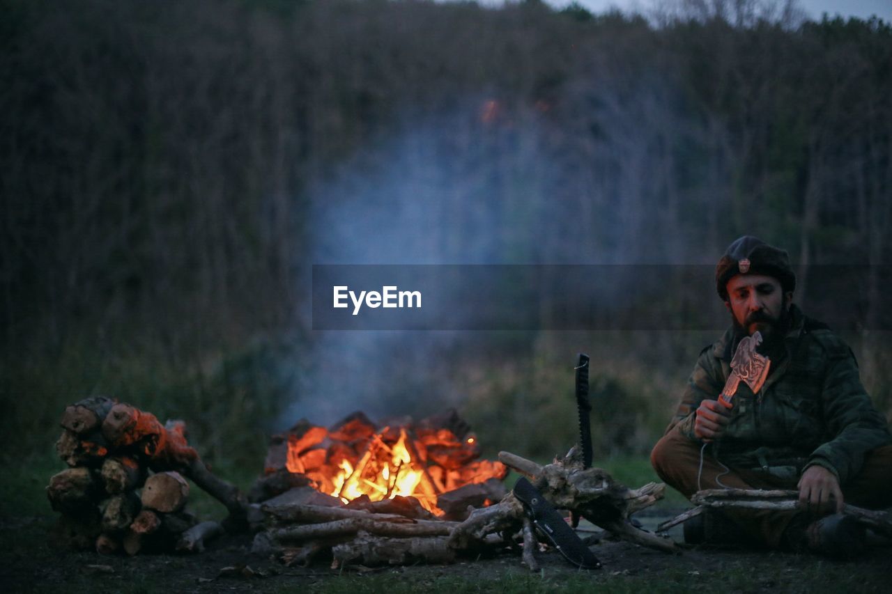 Mid adult man cutting wood by campfire on field at dusk