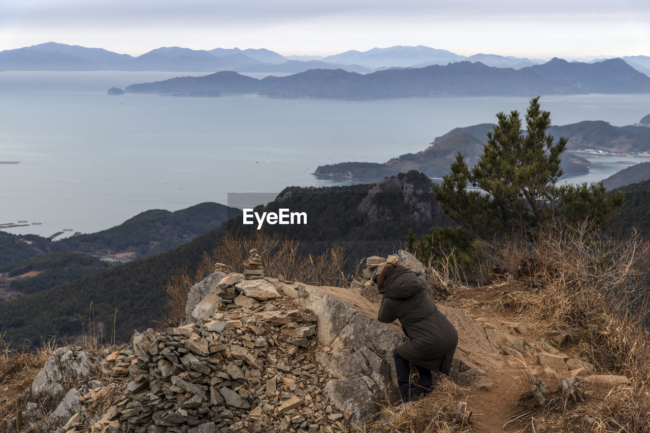 Side view of mature woman photographing sea while crouching on mountain against sky