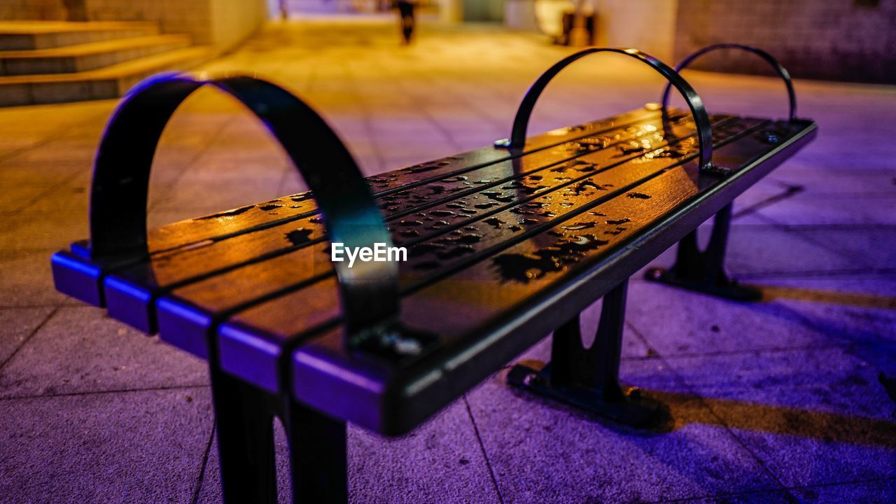 Close-up of wet metallic bench on sidewalk in city