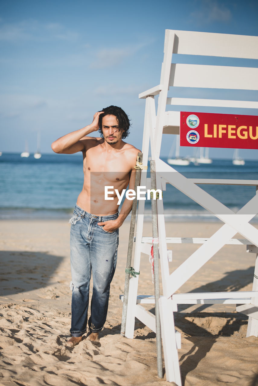 PORTRAIT OF SHIRTLESS MAN STANDING AT BEACH