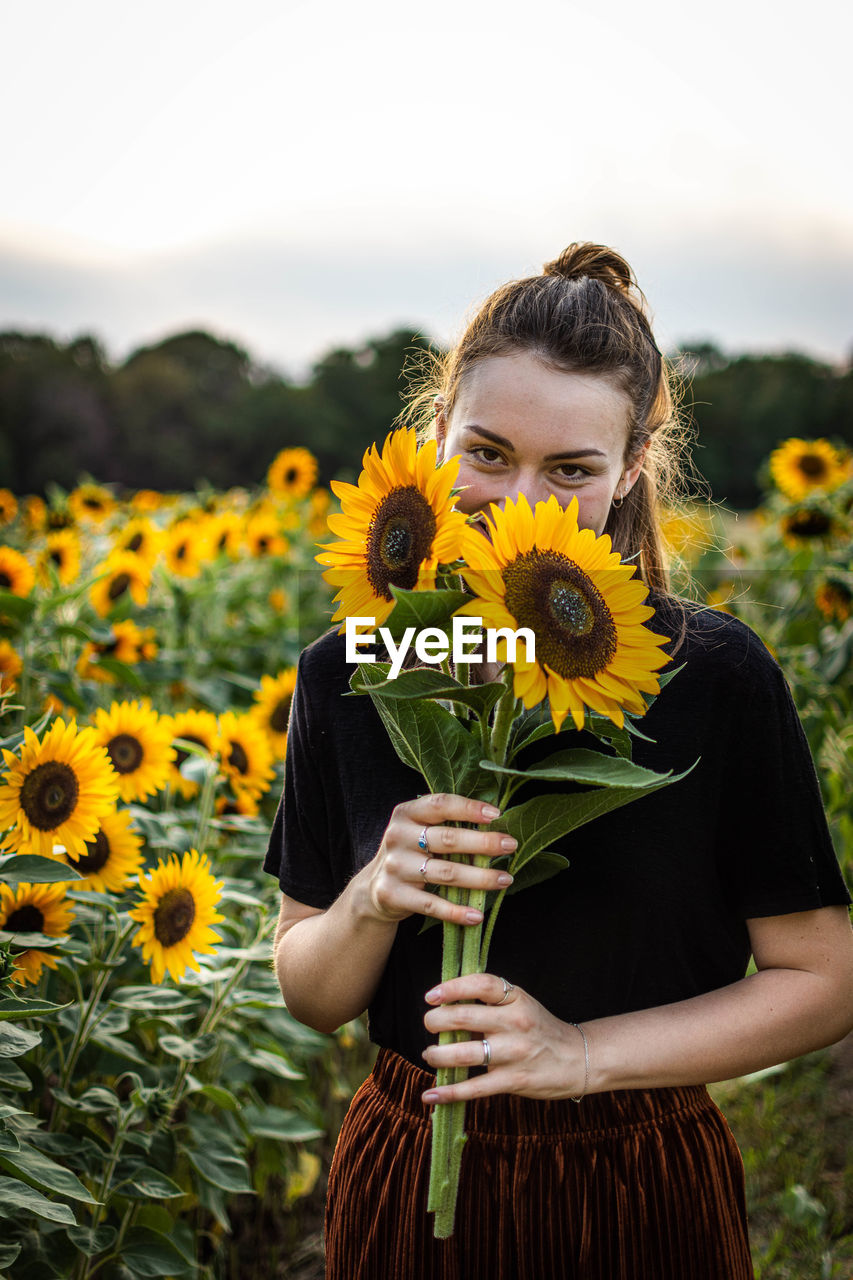 PORTRAIT OF WOMAN WITH SUNFLOWERS ON FIELD AGAINST ORANGE BACKGROUND