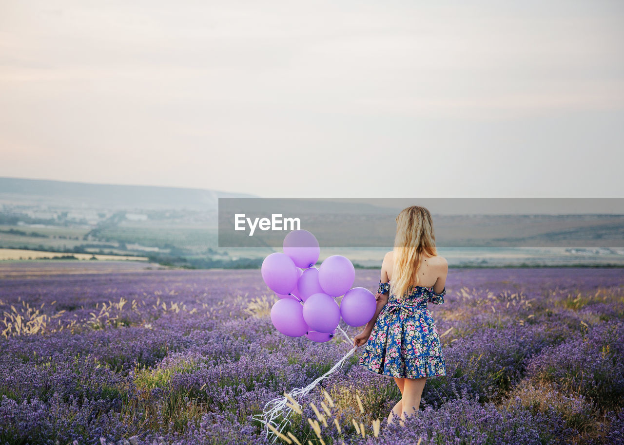 REAR VIEW OF WOMAN STANDING ON FIELD BY PURPLE FLOWERS