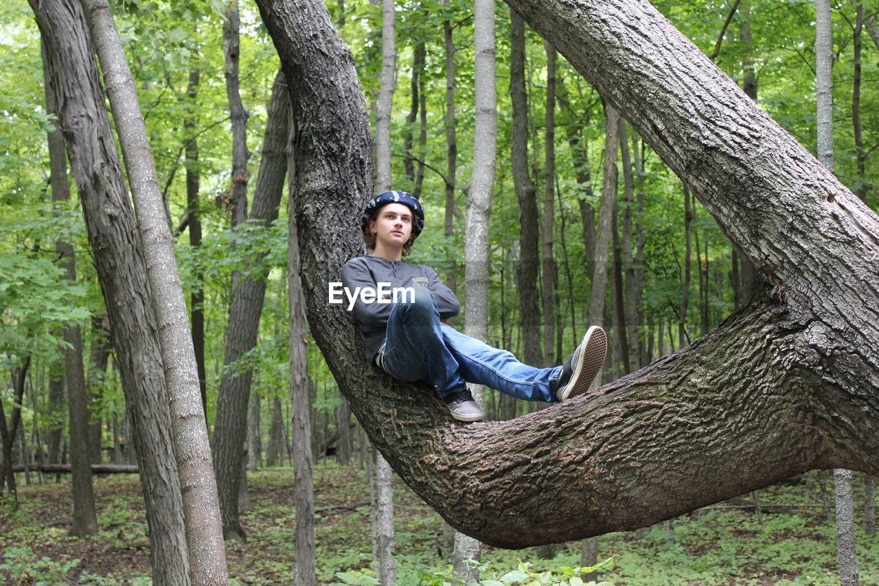 Full length of thoughtful young man sitting on tree trunk in forest