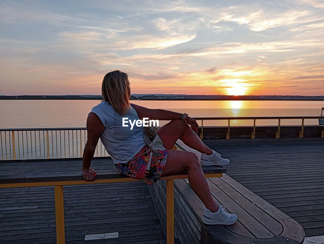 sky, sunset, water, one person, women, relaxation, adult, sea, lifestyles, nature, railing, leisure activity, beauty in nature, vacation, trip, holiday, full length, tranquility, cloud, sitting, dusk, young adult, casual clothing, summer, tranquil scene, female, scenics - nature, architecture, travel, travel destinations, sun, sunlight, land, rear view, idyllic, twilight, ocean, outdoors, beach, looking at view, hairstyle, horizon, horizon over water, tourism, pier, activity, copy space, enjoyment, long hair, blue, person