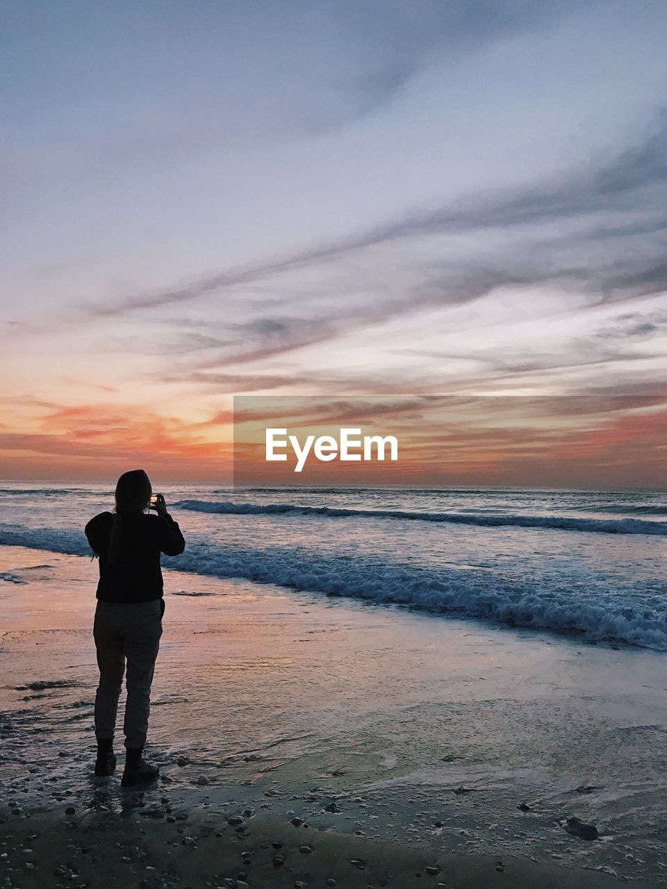 REAR VIEW OF COUPLE STANDING AT BEACH DURING SUNSET