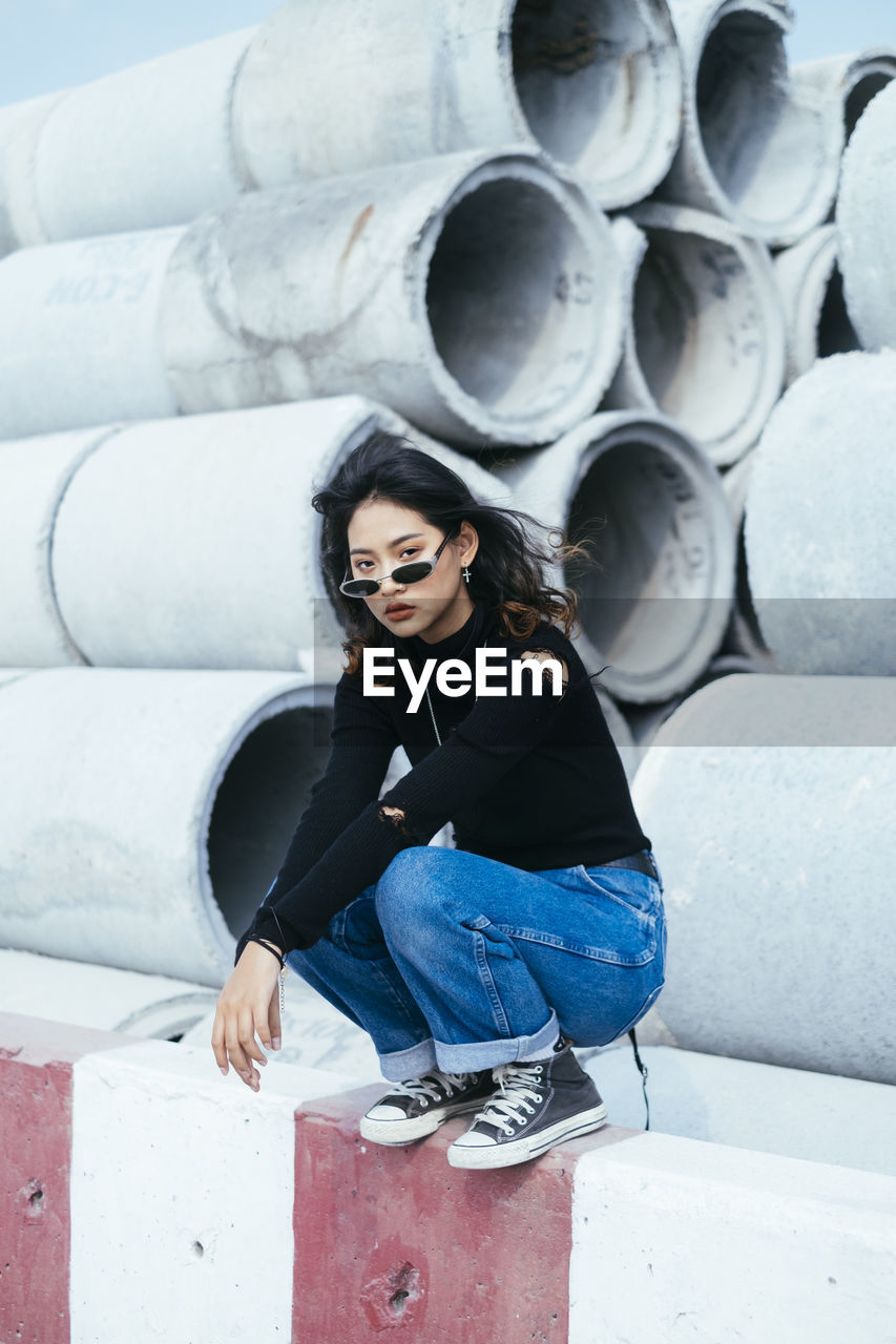 PORTRAIT OF YOUNG WOMAN WEARING SUNGLASSES STANDING AGAINST WALL