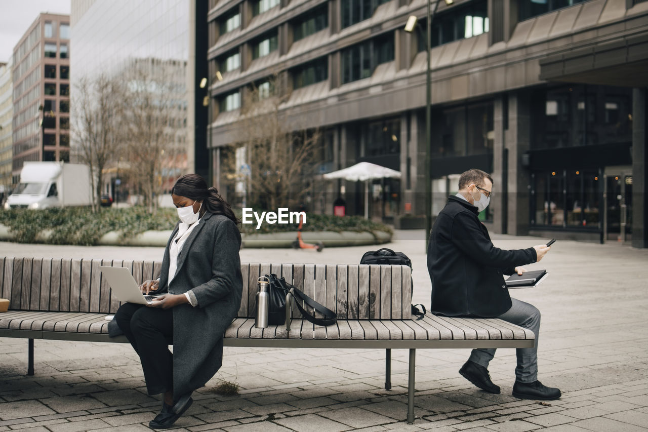 Businessman with smart phone while female colleague using laptop sitting on bench during covid-19