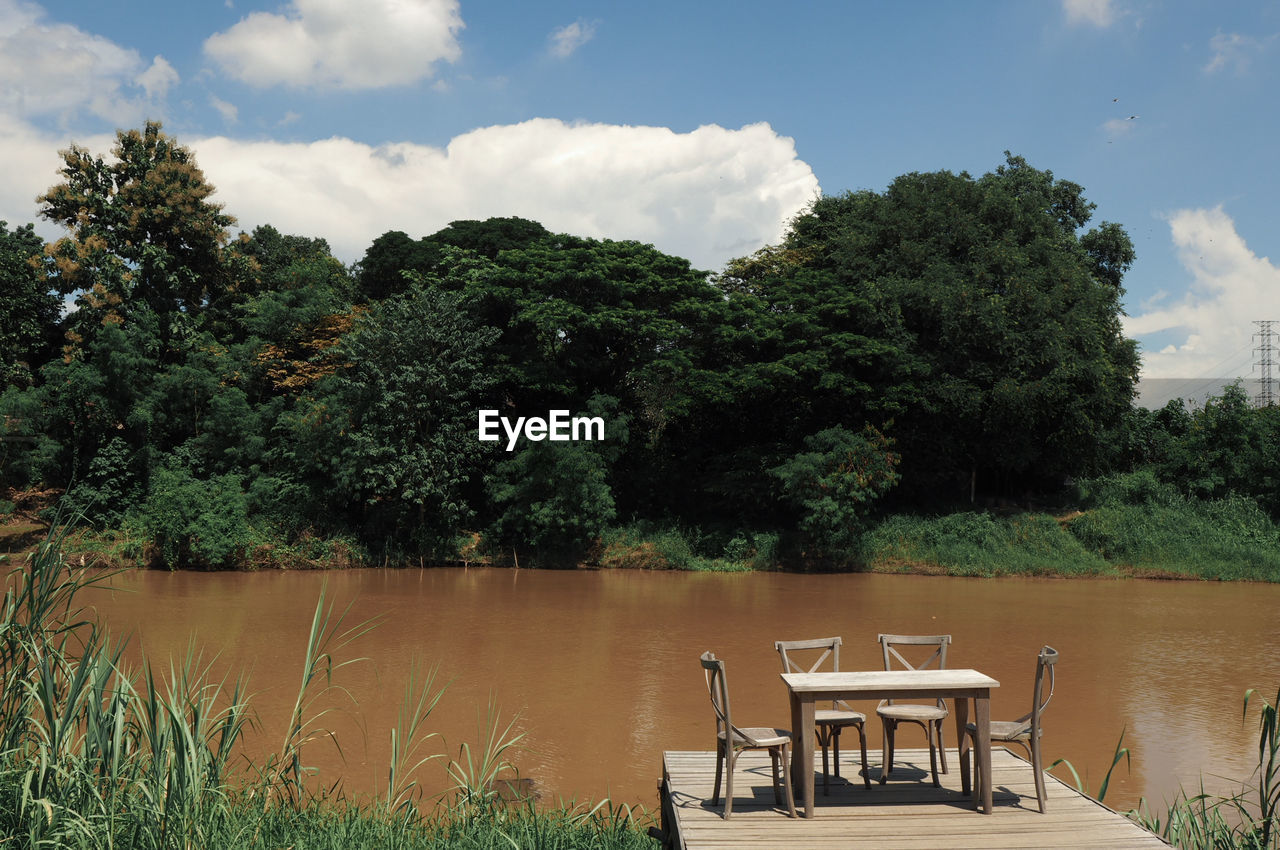 Empty chairs and tables by lake against sky during sunny day