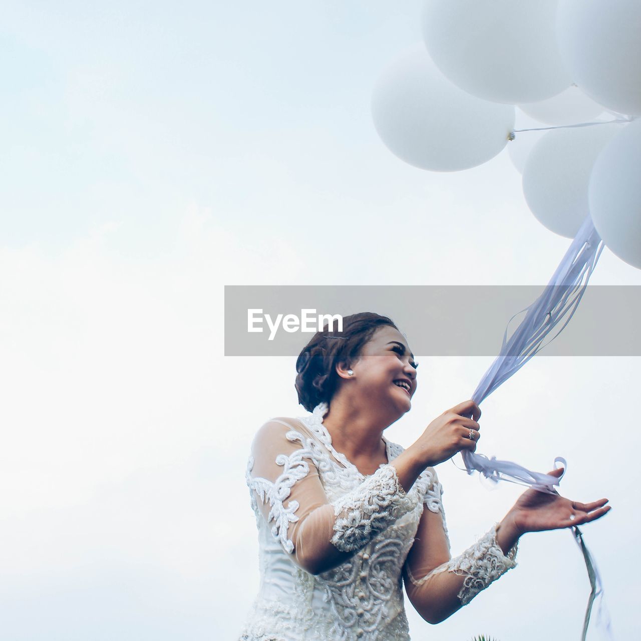 Cheerful bride holding helium balloons against sky