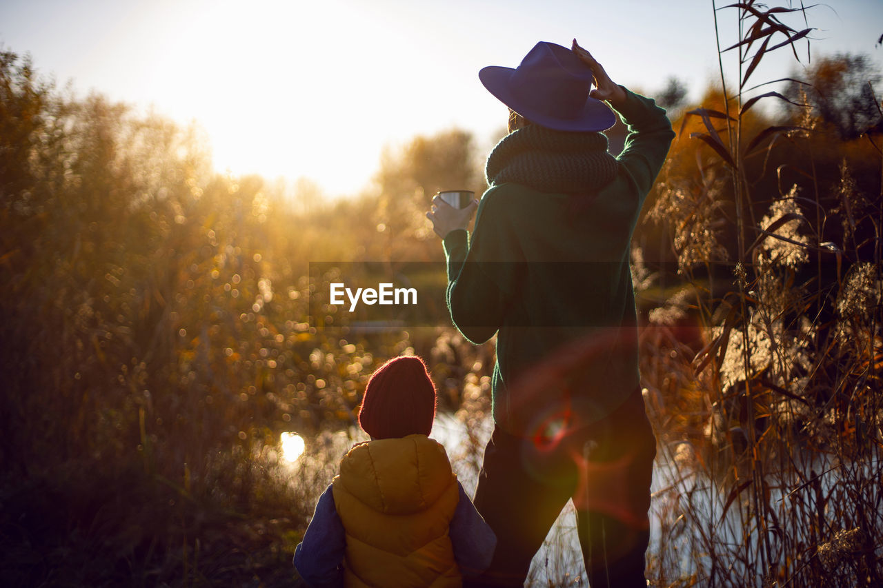 Woman in a hat and a boy stand with their backs to the lake at sunset in autumn