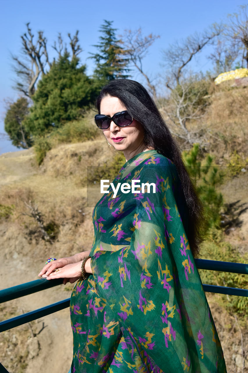 fashion, sunglasses, one person, glasses, adult, clothing, dress, women, nature, young adult, spring, portrait, photo shoot, green, land, blue, plant, standing, sky, smiling, three quarter length, tree, day, leisure activity, hairstyle, outdoors, looking at camera, lifestyles, sunlight, sunny, happiness, traditional clothing, landscape, travel, waist up, multi colored, female, brown hair, pattern, long hair, clear sky, emotion, travel destinations, arts culture and entertainment