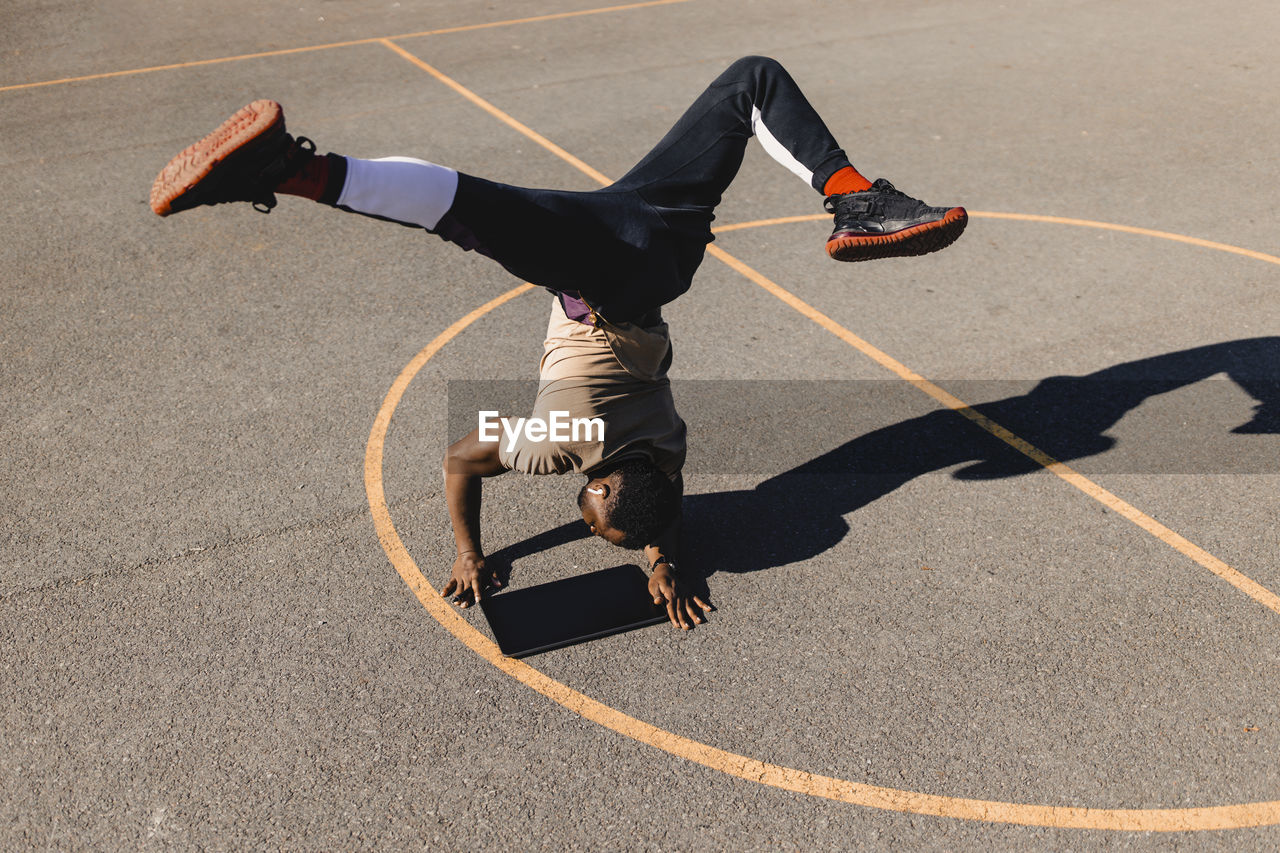 Young man balancing with handstand using graphics tablet on basketball court during sunny day