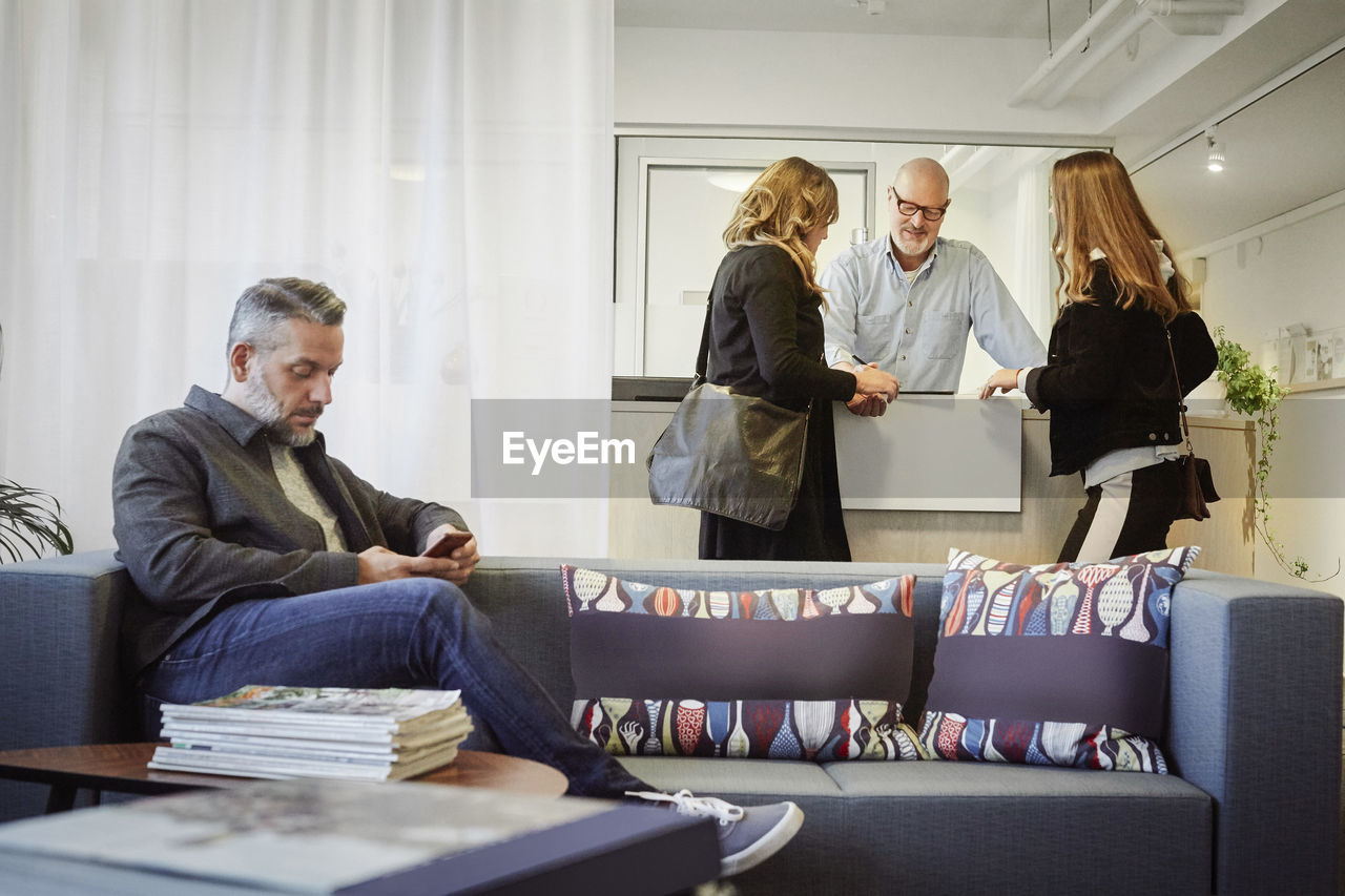 Mother and daughter talking to male therapist at reception while man using phone in waiting area