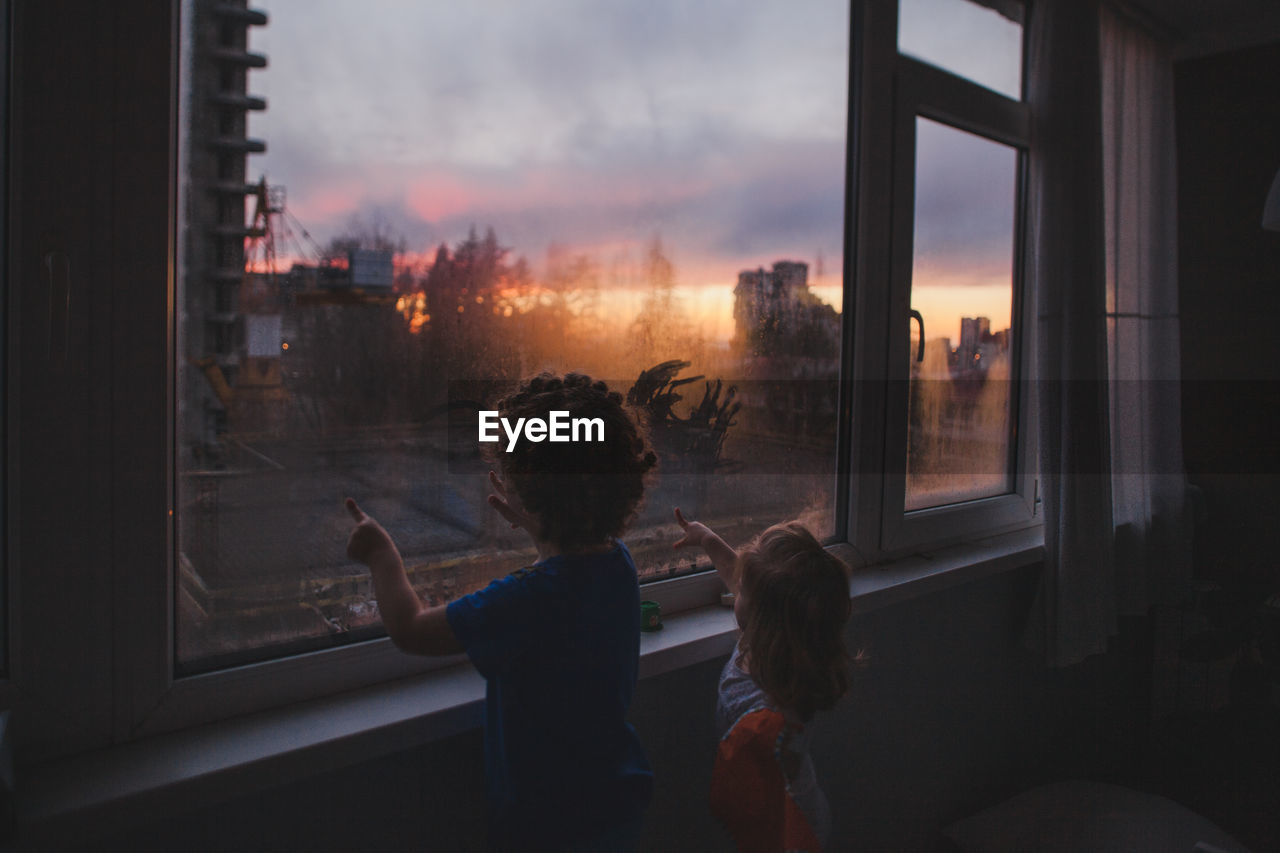 Children sit at home with their fingers drawing on the window at sunse