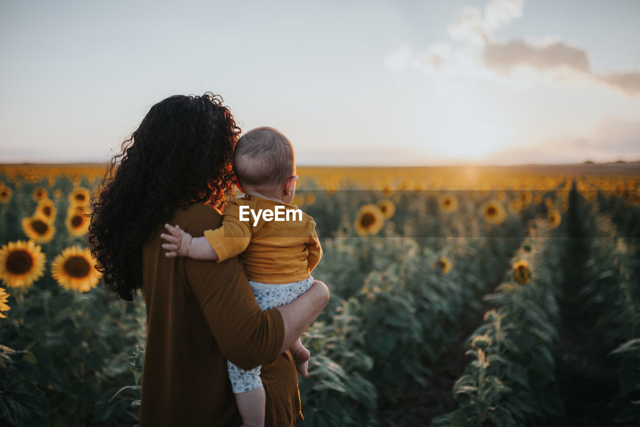 Mother carrying toddler daughter in sunflower farm against sky