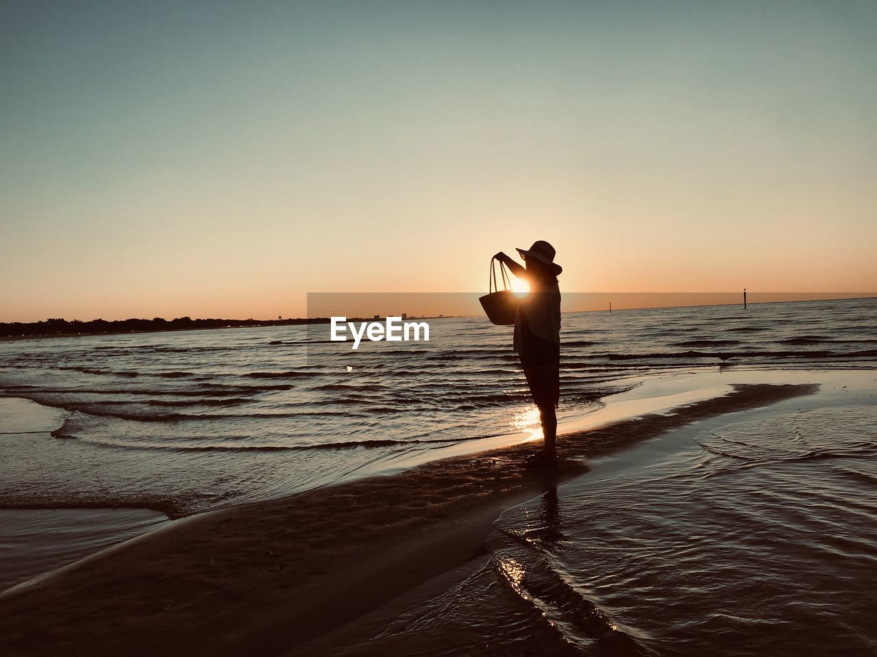 Silhouette woman holding bag standing on beach during sunset