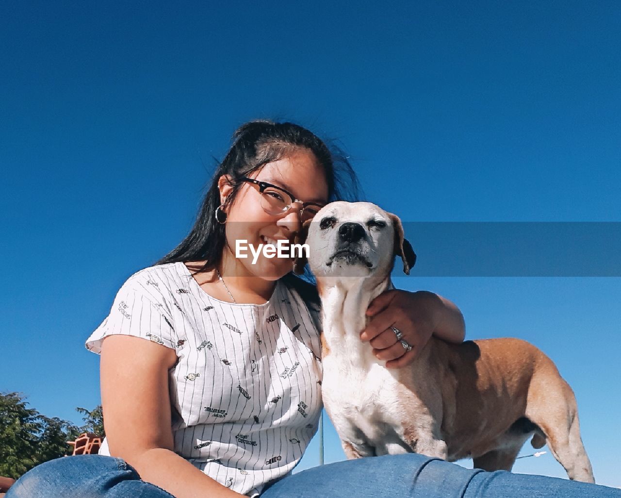 Portrait of smiling young woman with dog sitting against blue sky during sunny day