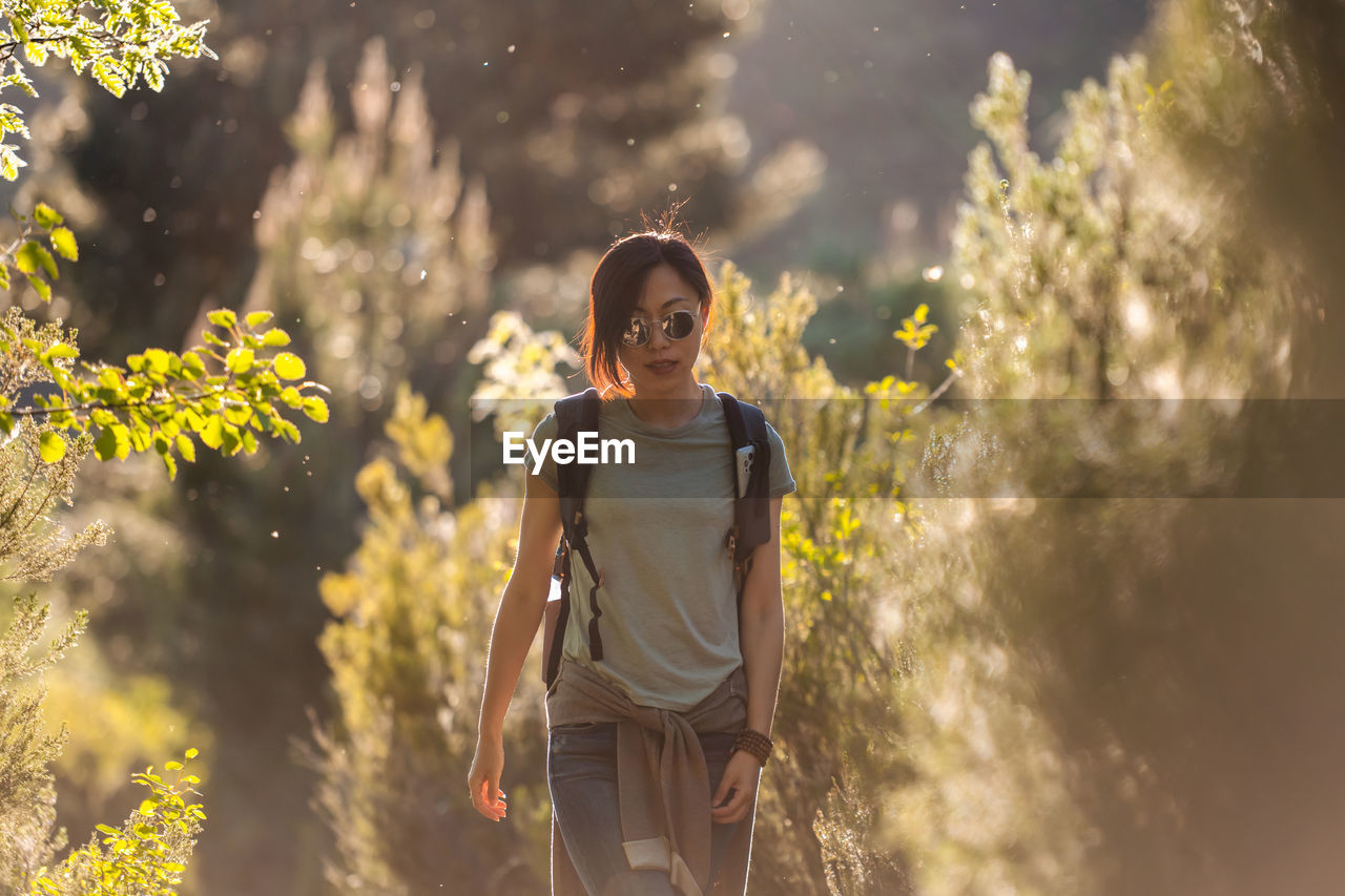 Asian female hiker in casual wear and sunglasses with rucksack strolling in nature with green plants during trekking on sunny summer day in valverde de los arroyos