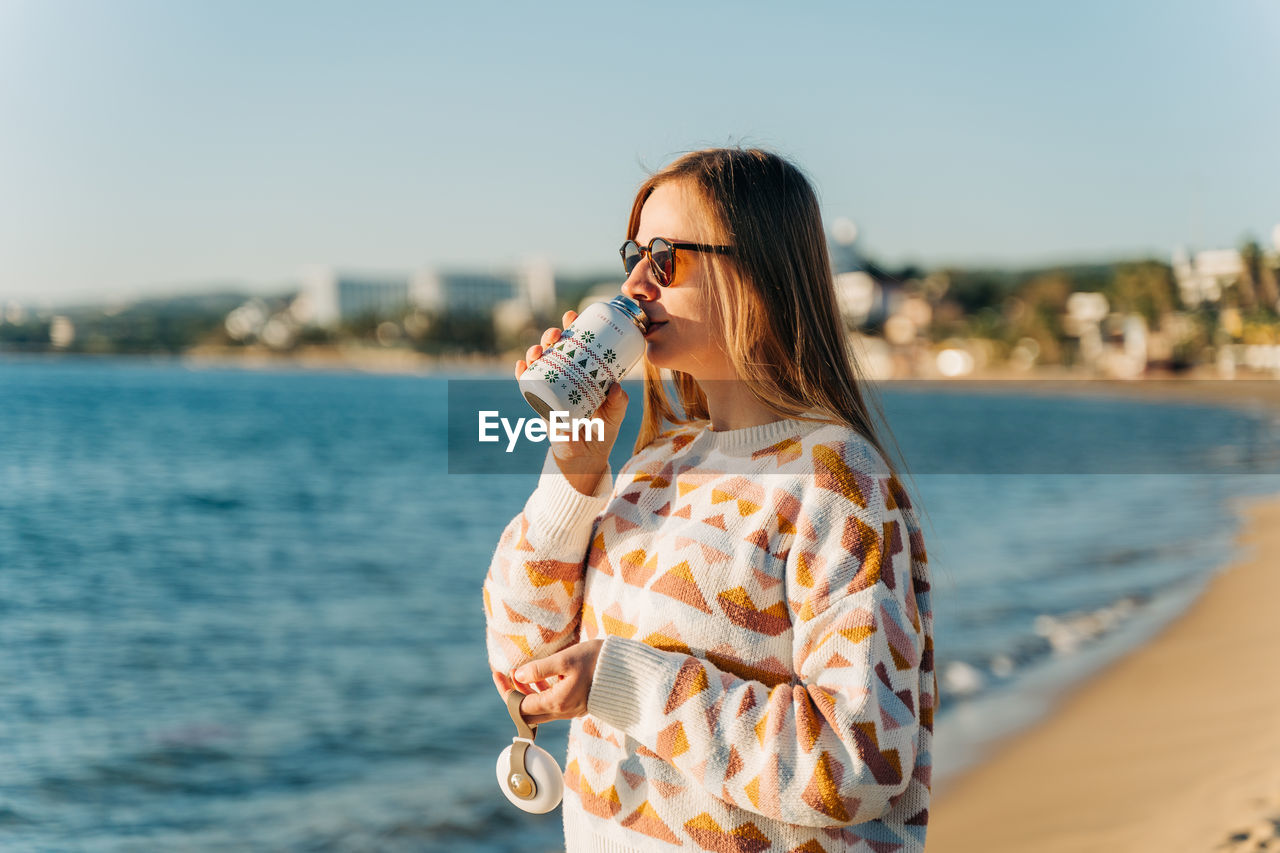 young woman drinking water while standing against sea against sky