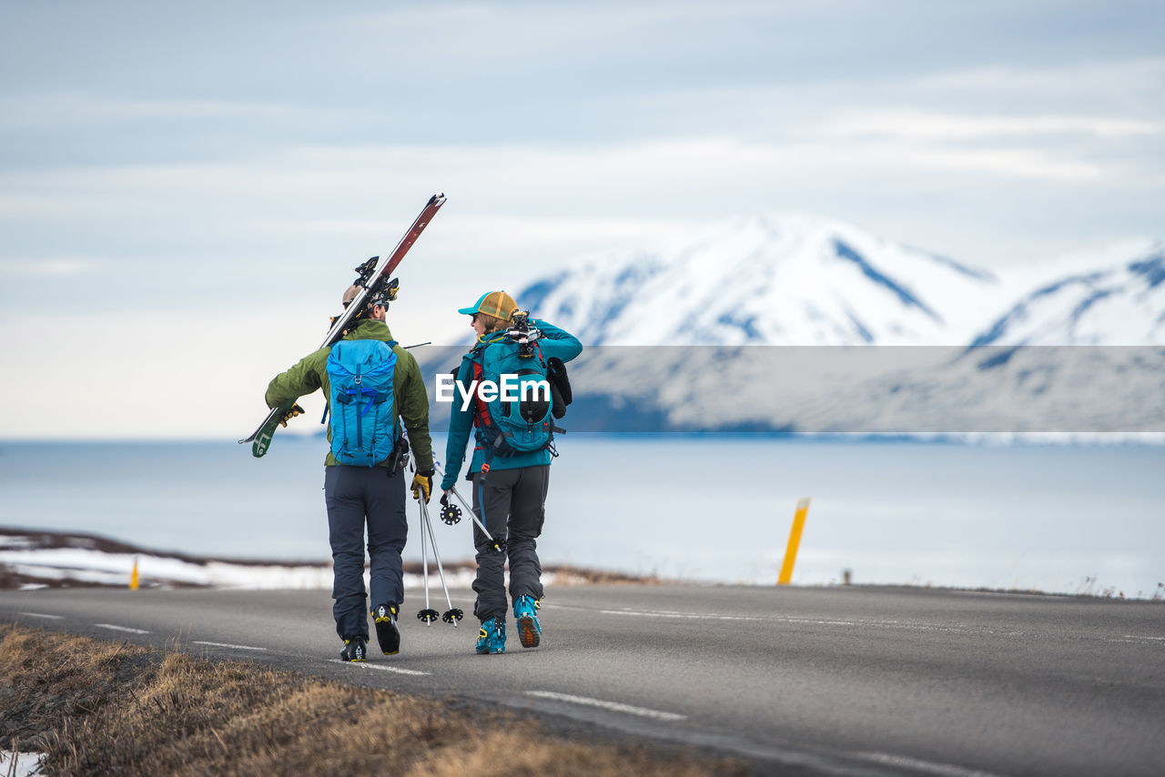 A couple walks down a road in iceland holding ski gear