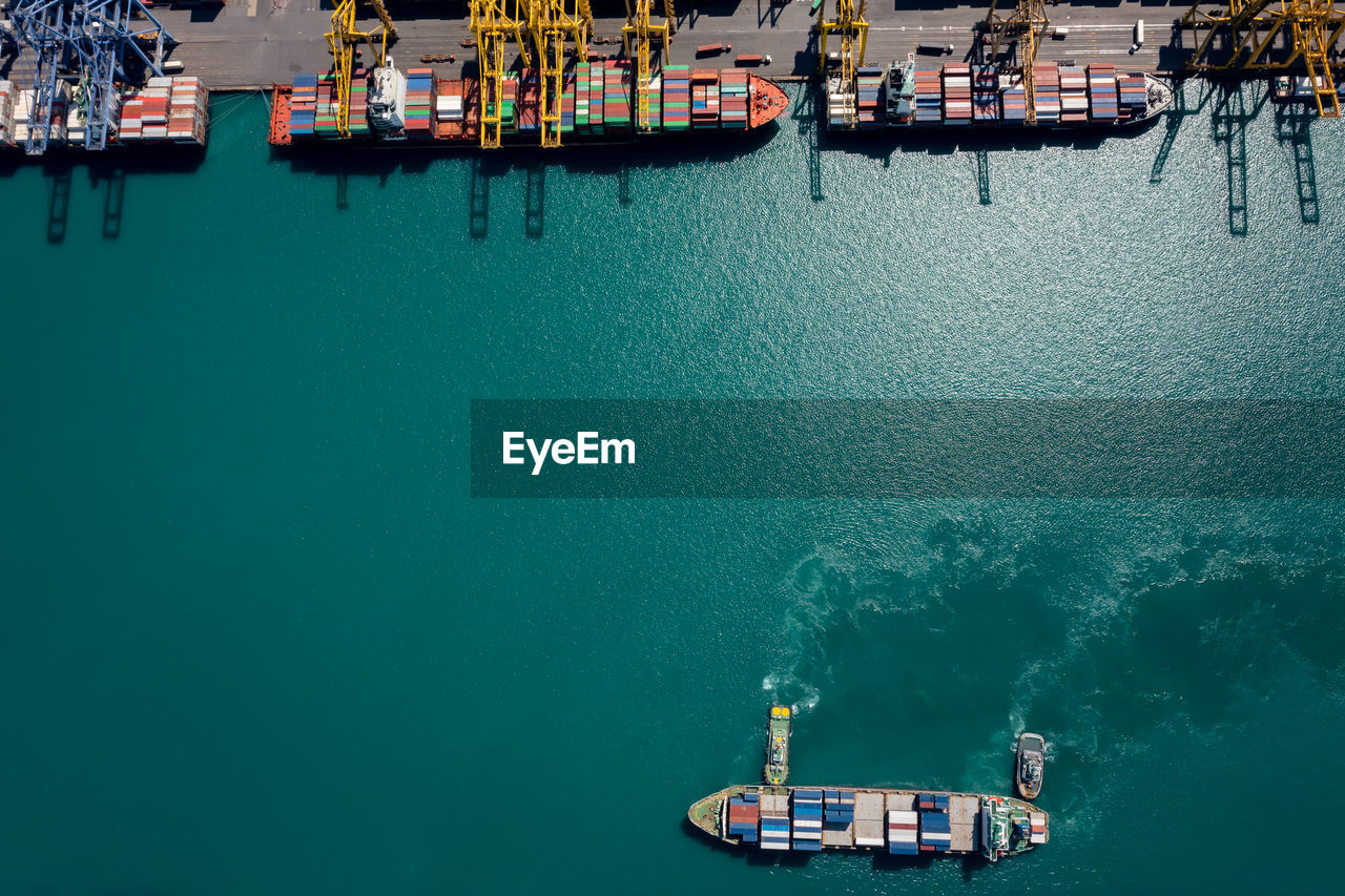 Aerial view container ship and tugboat sailing in sea and commercial port working crane bridge 