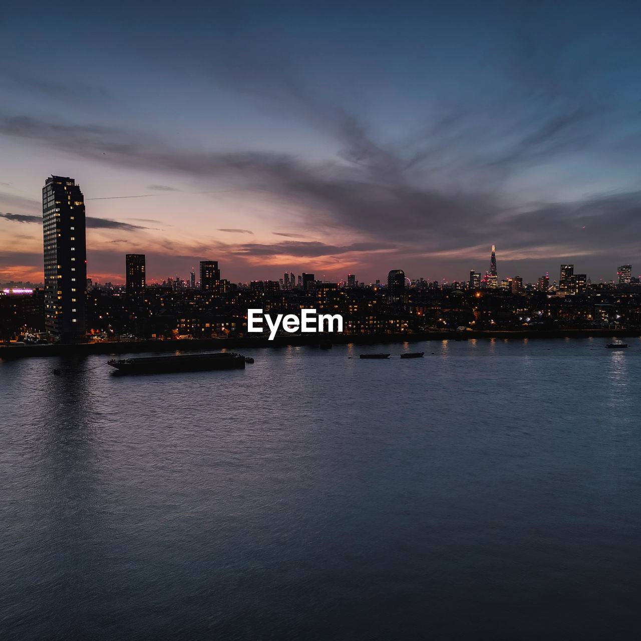 Illuminated buildings by river against sky at sunset