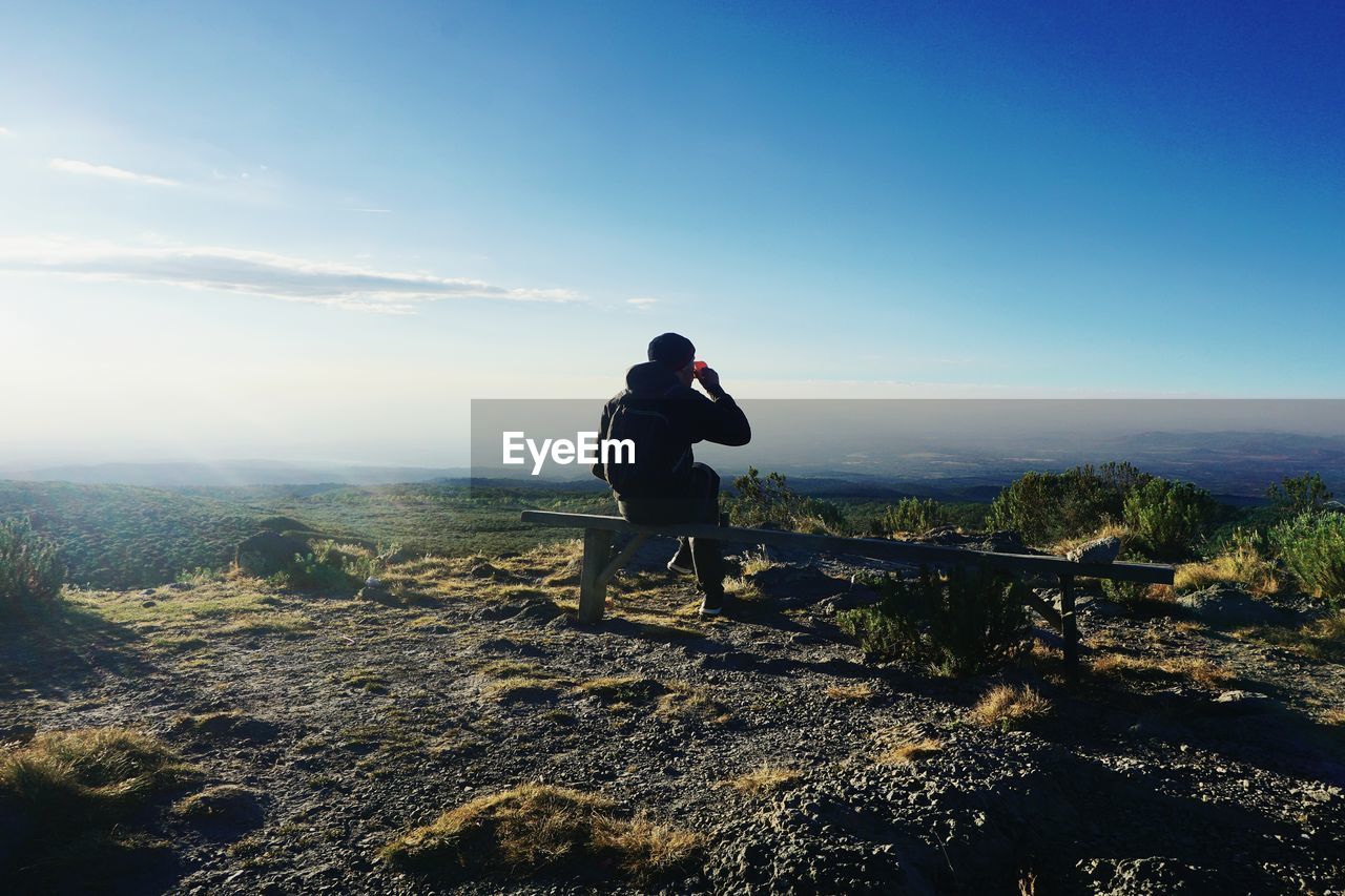 Rear view of man having drink while sitting on bench at mountain against sky
