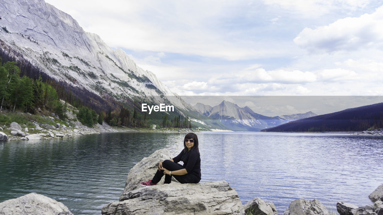 WOMAN SITTING ON ROCK BY LAKE AGAINST MOUNTAINS