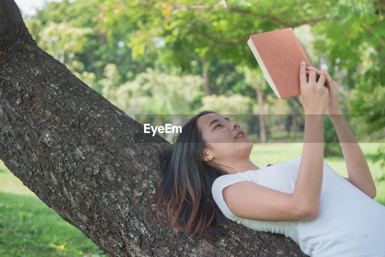 YOUNG WOMAN USING PHONE WHILE SITTING ON TREE