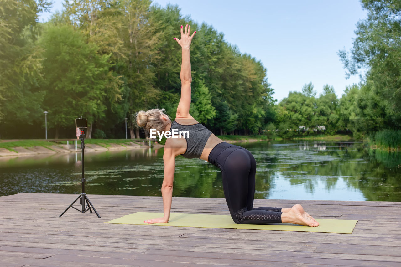 A woman in a on a wooden platform in summer, does yoga by pond in park