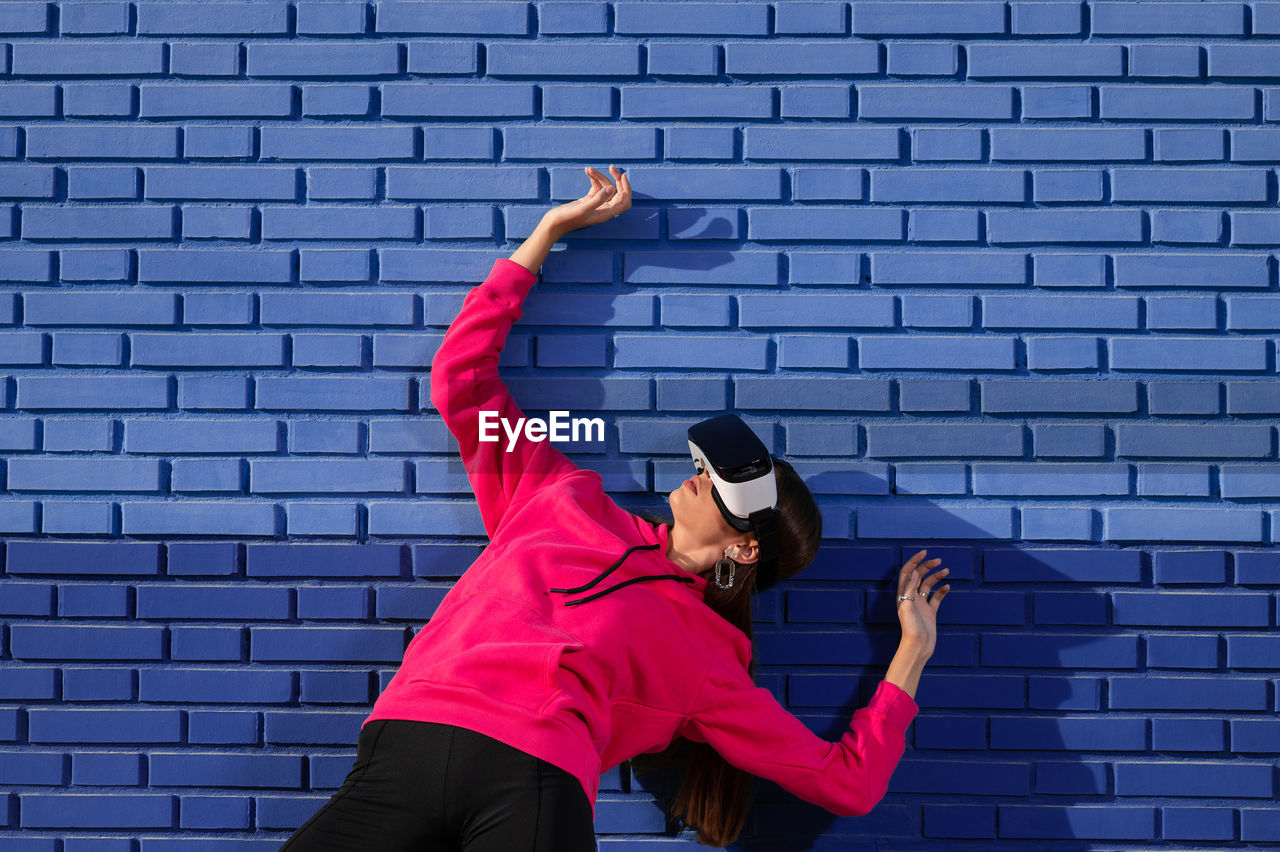 one person, adult, brick wall, clothing, sports, brick, arms raised, arm, wall, lifestyles, young adult, sports clothing, dancing, wall - building feature, limb, human limb, vitality, exercising, red, architecture, leisure activity, person, motion, strength