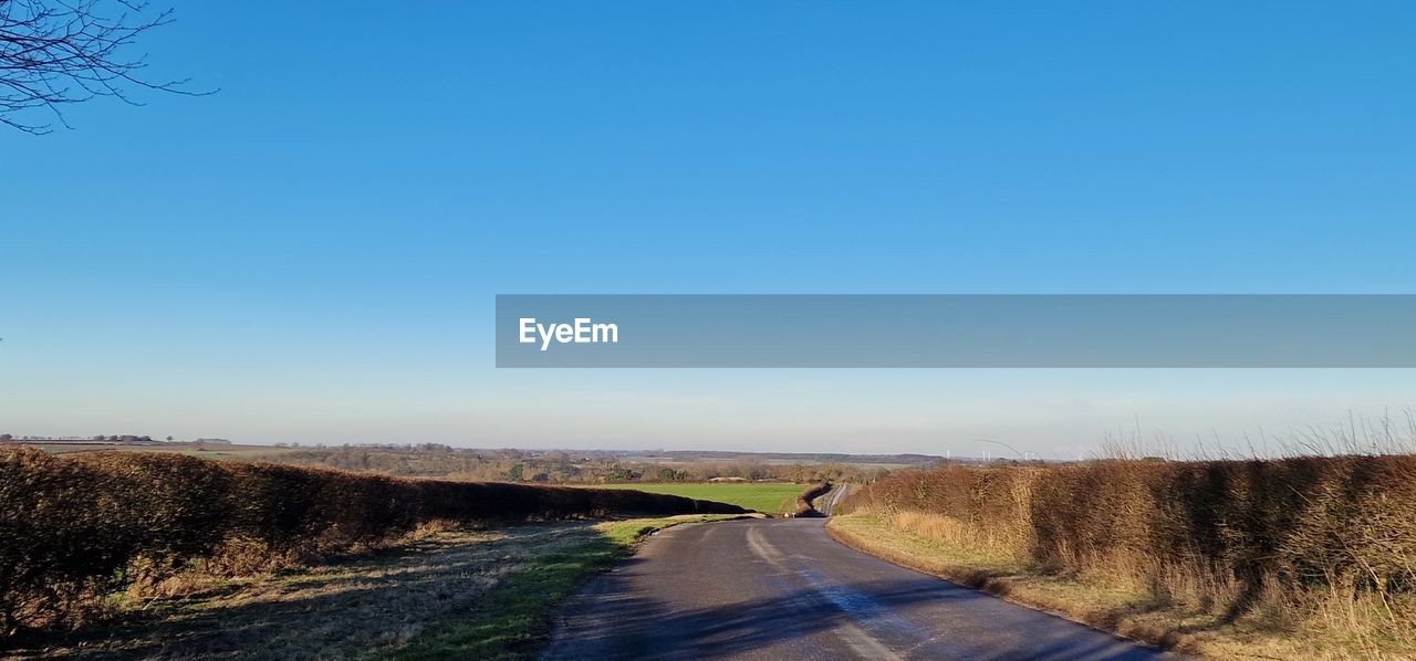 sky, road, the way forward, landscape, nature, hill, transportation, blue, clear sky, plant, environment, rural area, horizon, scenics - nature, no people, infrastructure, tranquility, beauty in nature, morning, country road, diminishing perspective, grass, tranquil scene, tree, land, copy space, non-urban scene, day, vanishing point, empty road, sunlight, rural scene, prairie, sunny, field, footpath, dirt road, outdoors, dirt, highway, travel, natural environment, remote, idyllic, road trip, horizon over land