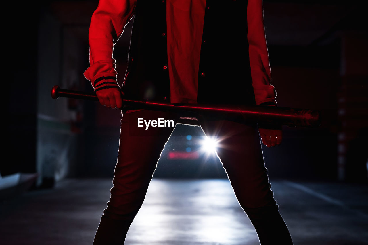 Crop legs of faceless woman in jeans and black boots with massive sole holding bat with car headlights on background