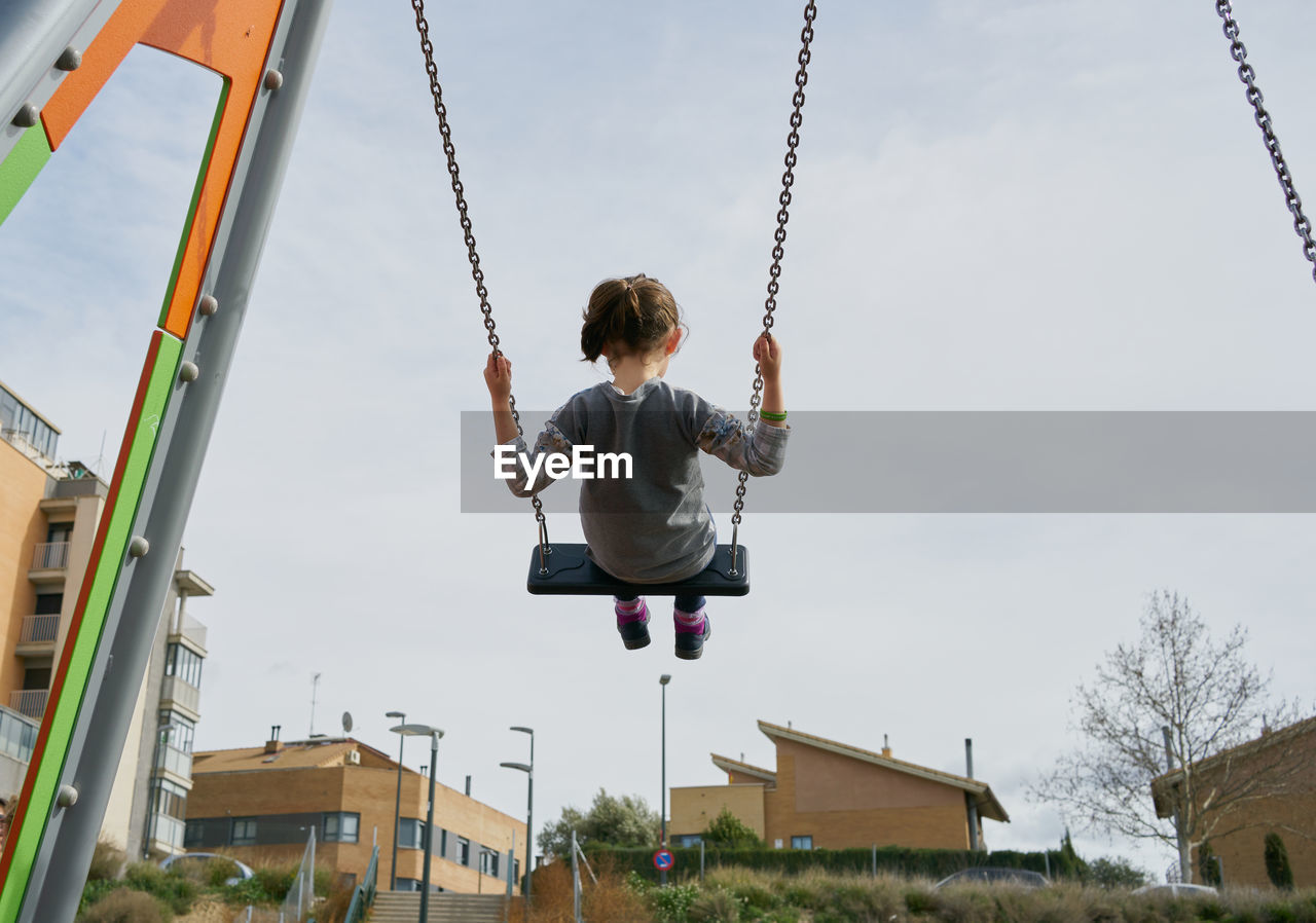 Back view of happy little girl swinging high on playground swing set