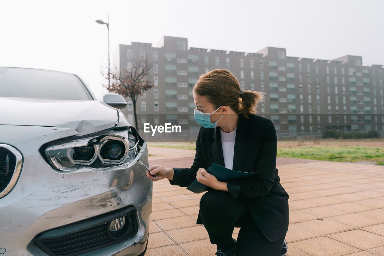 Side view of unrecognizable female insurance agent in formal outfit and protective mask writing protocol near damaged car parked on street after accident