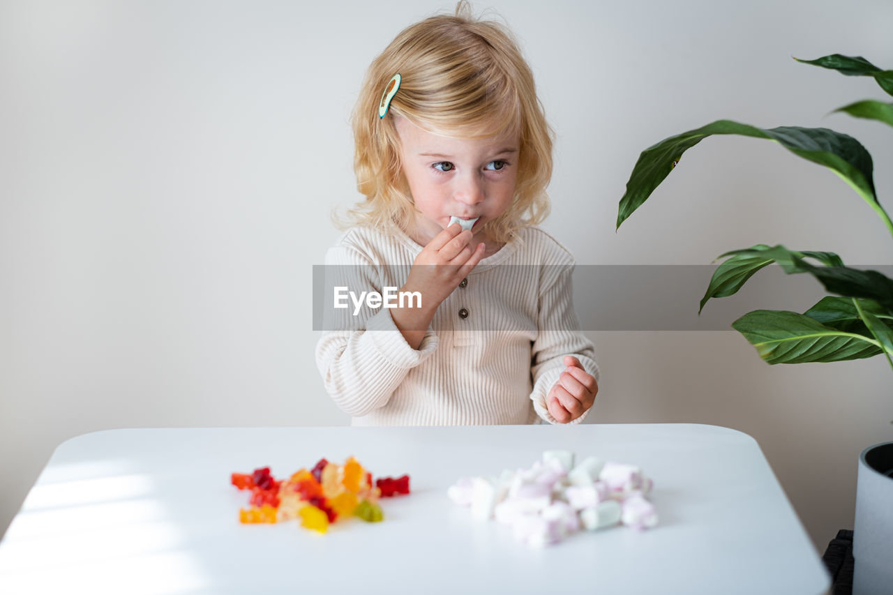 Cute caucasian blonde curly-haired baby girl two, three year old eating jelly, sweet gummy, candy