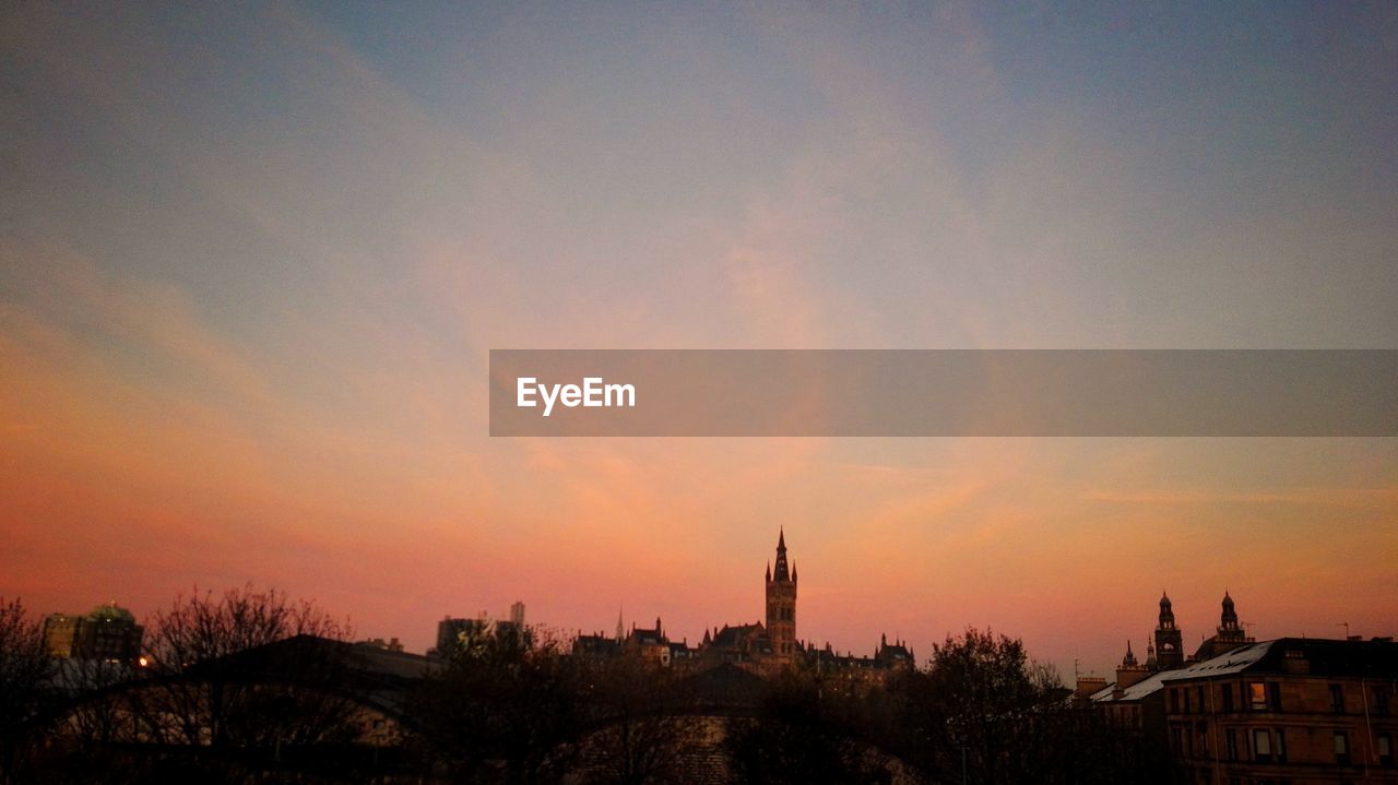 Buildings by trees against cloudy sky during sunset