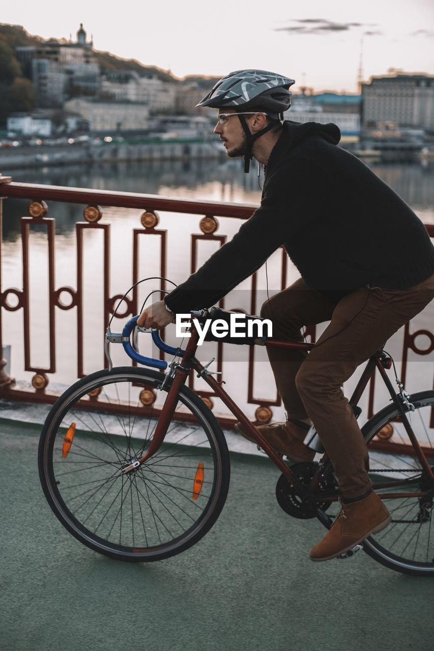 Side view of male cyclist riding bicycle on bridge over river in city during sunset
